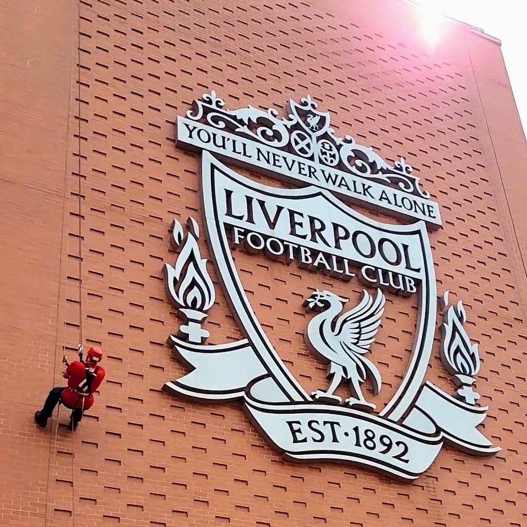 Man abseiling down the side of Anfield stadium next to the LFC crest.